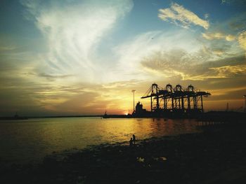 Silhouette cranes at harbor in sea against sky during sunset