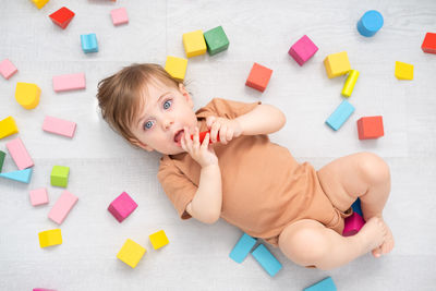Cute smiling baby girl in brown bodysuit laying on floor around colorful wooden blocks. top view
