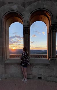 Woman standing by window against sky during sunset