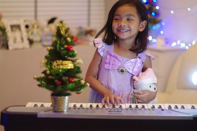 Cute girl holding stuffed toy while playing piano by christmas tree