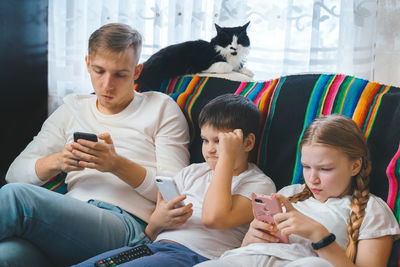 Siblings using smart phones while sitting on sofa with cat at home