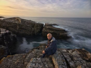 Portrait of senior man sitting on rock by sea against sky during sunset