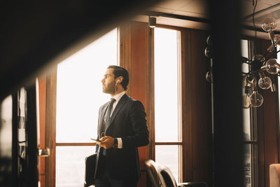 Thoughtful male financial advisor with headphones talking on mobile phone while standing by window at office