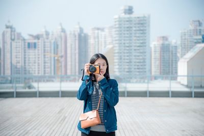 Portrait of young woman photographing against city