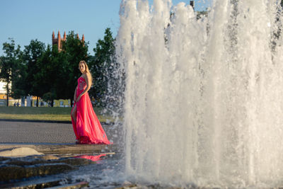 Young woman in pink evening gown standing by fountain in park