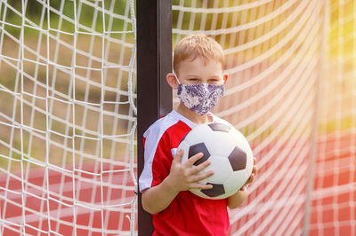 Portrait of boy wearing mask holding soccer ball standing by net
