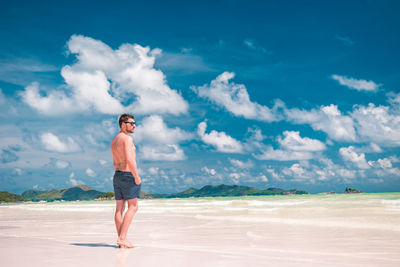 Shirtless mid adult man standing at beach against cloudy sky
