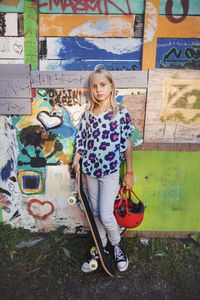 Portrait of girl holding skateboard and helmet while standing against graffiti wall