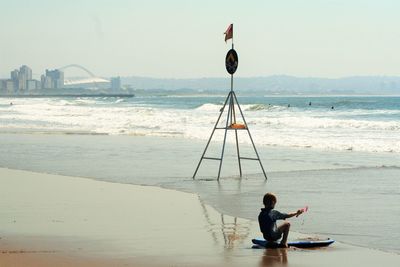 Playful girl sitting on surfboard by sea against clear sky