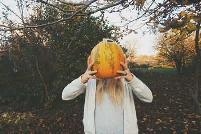 Woman covering face with pumpkin while standing by autumn trees on field