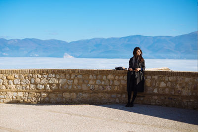 Full length of woman leaning on retaining wall by mountains against clear blue sky