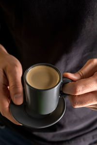A man's hand holds a cup of hot espresso coffee against the background of a black t-shirt.