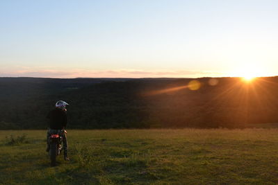 Rear view of man with motorcycle on grassy field against sky during sunset