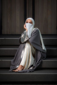 Full length image of veiled woman with white niqab sitting on the stairs and looking at the camera