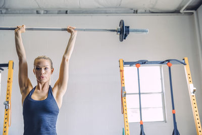 Confident young woman lifting barbell against wall in health club