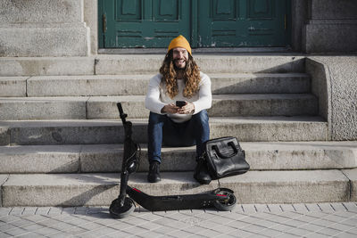 Laughing male hipster with mobile phone sitting on steps during sunny day