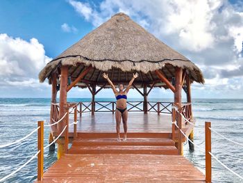 Portrait of smiling bikini woman with arms raised standing on gazebo over sea