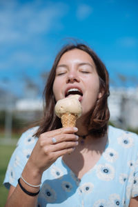 Young woman with eyes closed eating ice cream cone while standing against blue sky during sunny day