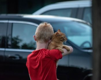 Rear view of boy with cat standing against cars