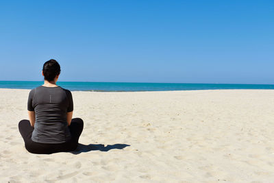 Rear view of man sitting on beach against clear sky