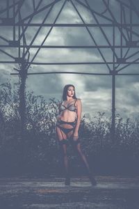 Young woman in lingerie standing at park against cloudy sky