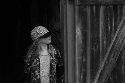 Girl wearing cap looking away while standing by wooden wall