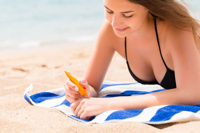 Smiling woman holding sunscreen bottle while lying on towel at beach