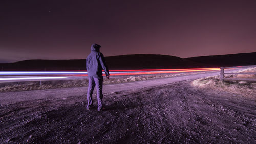 Rear view of man standing by light trails on road at night