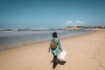 Rear view of woman with bucket walking at beach against sky