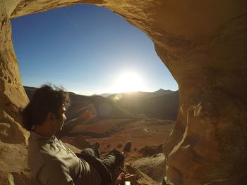 Close-up of man in cave by landscape against sky in sunny day