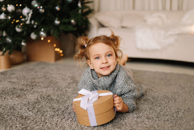 A little girl child in a knitted sweater holds a christmas gift box on holiday in a decorated house