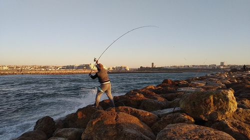 Man fishing on rock by sea against clear sky