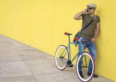 Man talking on phone while standing with bicycle against yellow wall