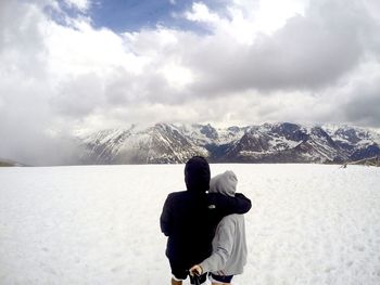 Rear view of couple photographing on snow covered field by mountains against cloudy sky