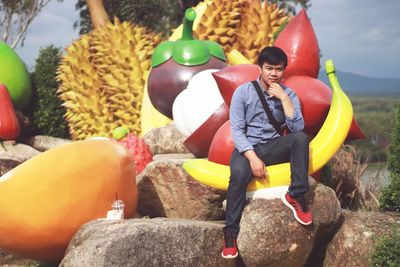 Full length portrait of man sitting on rock against artificial fruits