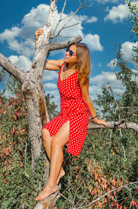 Young woman in a red dress in white polka dots sitting on a branch of dry tree in garden