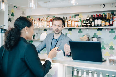 Owner looking at businesswoman doing contactless payment in office cafe