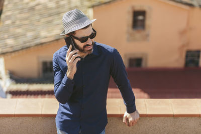 Attractive middle-aged man with hat and sunglasses talking to a smart phone standing on balcony.