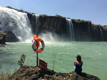 Rear view of woman sitting on rock against waterfall