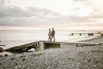 Couple walking on jetty at beach against sky during sunset