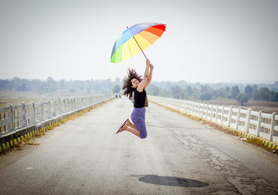 Full length of woman jumping with umbrella on road against clear sky
