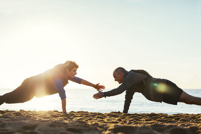 Side view of father and son doing push-ups together against clear sky at beach during sunset