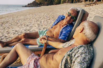Smiling gay man spending vacation with boyfriend reclining on deck chair at beach