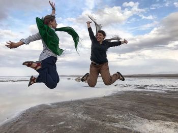 Full length of female friends jumping on shore against sky