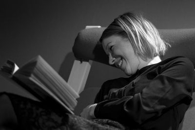 Close-up of smiling woman with book sitting on chair at home