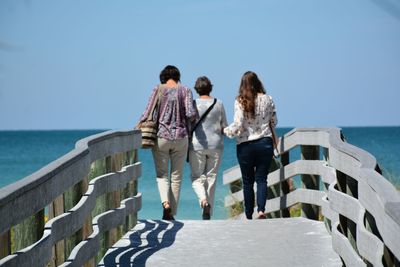Rear view of women walking on footbridge against sea and clear sky