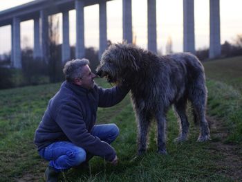 Mature man with dog on field against bridge