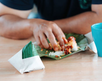 Nasi lemak wrapped in banana leaf with brown paper with people eating in the background.