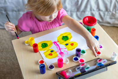 Little girl painting with paints, looking at tablet, sitting at the table at home. online school