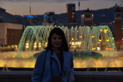 Portrait of smiling young woman standing against illuminated fountain at night